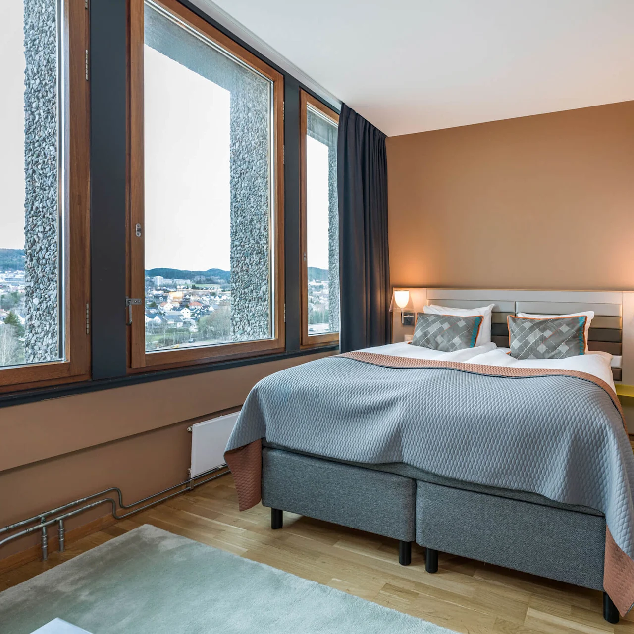 A neatly made double bed with gray linens sits in a warmly painted room, beside large windows overlooking a distant cityscape, enhancing the cozy, modern atmosphere.