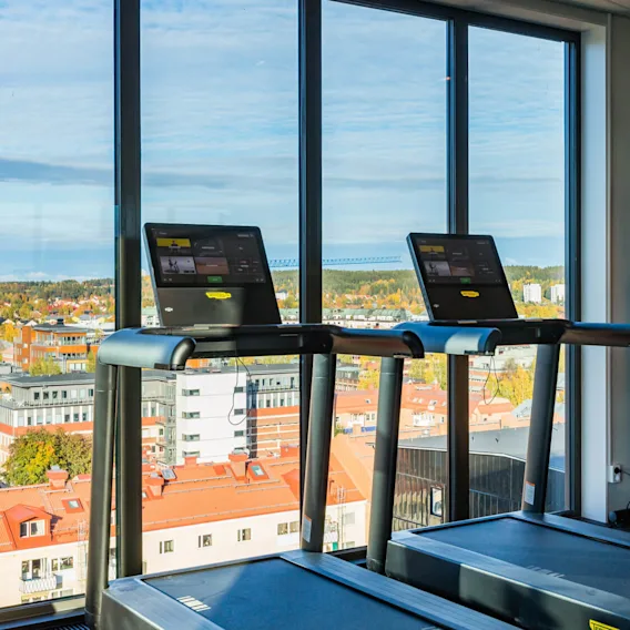 Två löpband med digitala skärmar står vid stora fönster med stadsutsikt i ett modernt gym. I närheten syns en multigymmaskin. Skylten lyder "Brandlarm".