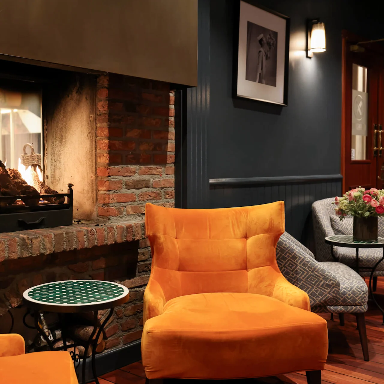 A cozy lounge with a burning fireplace set into a brick wall, surrounded by orange and patterned chairs, small tables, wood flooring, artwork, and dim lighting creating a warm atmosphere.