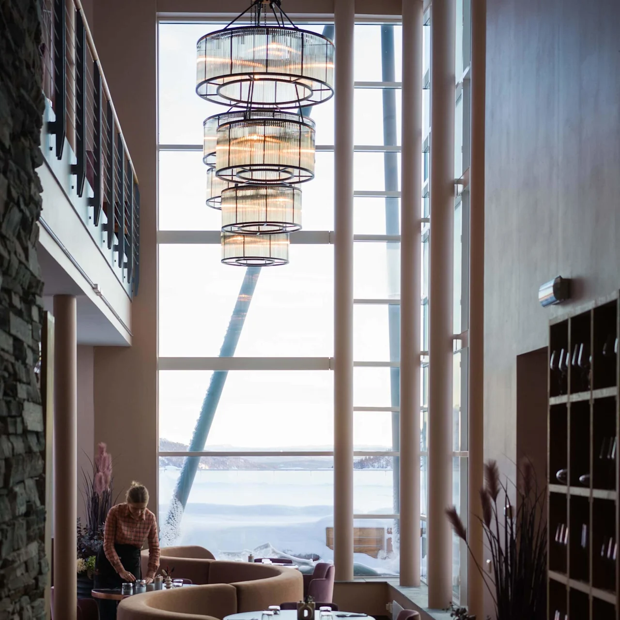 Elegant dining area with large windows, showcasing a snowy landscape. A person arranges items on a round table beneath a modern chandelier, surrounded by stylish furniture.