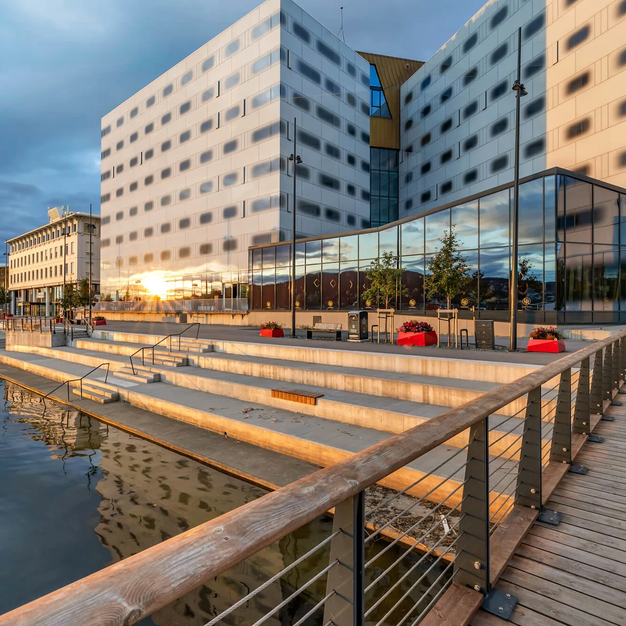 A modern building with reflective glass windows stands beside a stepped promenade by the water, under a cloudy sky. Red planters dot the area, creating a lively atmosphere.