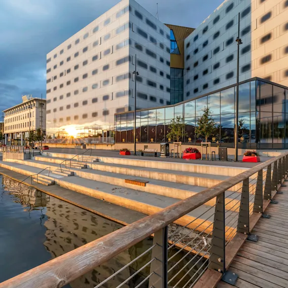 En moderne bygning med reflekterende glassvinduer står ved siden av en trappet promenade ved vannet, under en skyet himmel. Røde plantekasser piffer opp området.