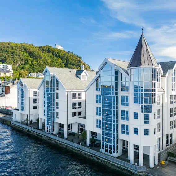 En moderne hvit bygning med store glassvinduer og et tårn, ved vannet med en grønn ås i bakgrunnen.
