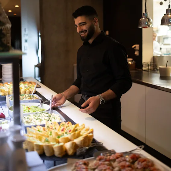 En smilende servitør i svart skjorte står bak et buffetbord fylt med ulike retter, salater og wraps.