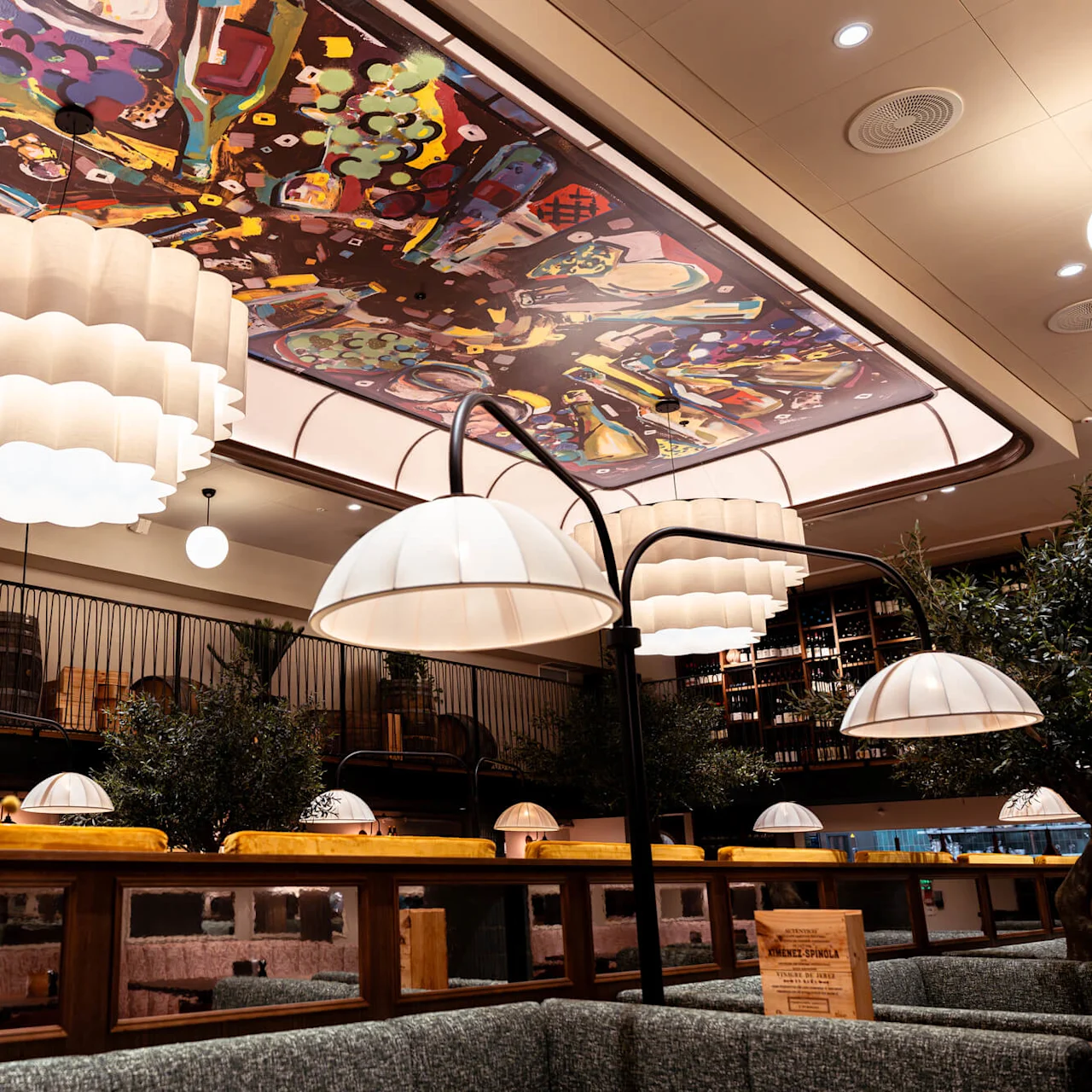 English: Elegant restaurant interior featuring large, ornamental overhead lamps illuminating tables below. Decorative ceiling art enhances ambiance, complemented by shelves of wine bottles and wooden barrels.