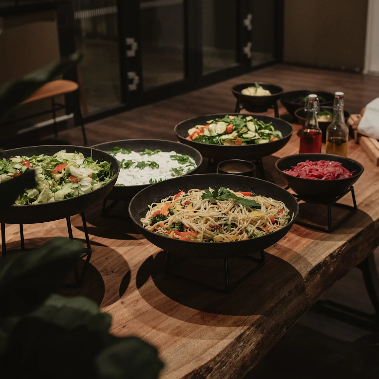 A wooden table holds plates of various salads and pasta dishes, accompanied by bread, oil, and vinegar bottles in a warmly lit dining room.