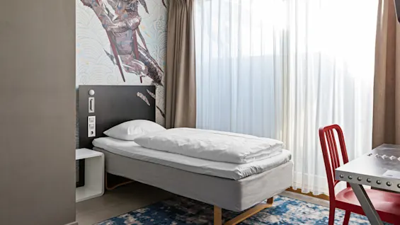 Single bed with white bedding against a mural wall, next to a large window with sheer curtains. A red chair is at a metallic desk.