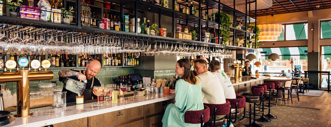 Bartender som förbereder drinkar bakom en lång, träbar. Gäster sitter på höga pallar och pratar med varandra. Hyllor med flaskor och glas hänger ovanför i en ljus, modern barmiljö.