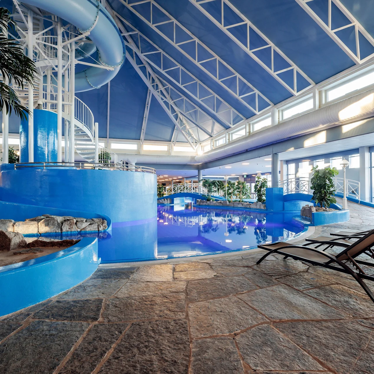Indoor pool area with a large spiral waterslide descending into the pool. Lounge chairs and tropical plants surround the area, set under a high, glass-paneled roof letting in natural light.