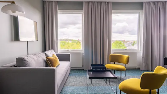 En grå sofa med gule puter står i en moderne stue. To gule stoler vender mot et svart kaffebord. Store vinduer med gardiner viser en skyet himmel.