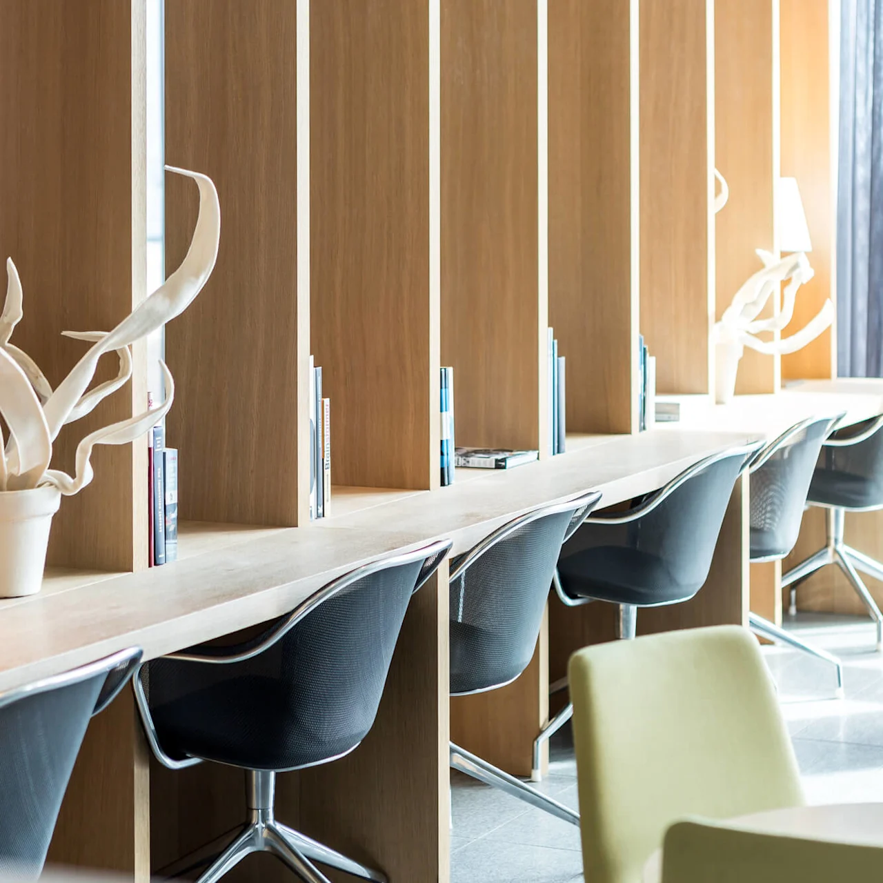Wooden cubicles with swivel chairs and books line a modern, well-lit workspace. Potted plants and lamps add decoration, while more seating is available with round tables and colorful chairs.