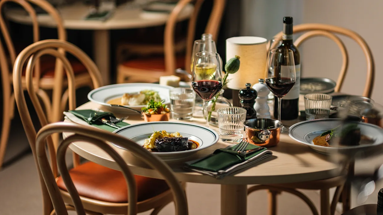 Ett runt matbord har tallrikar med gourmetmat, vinglas och en flaska, omgiven av trästolar i en varmt upplyst restaurangmiljö.
