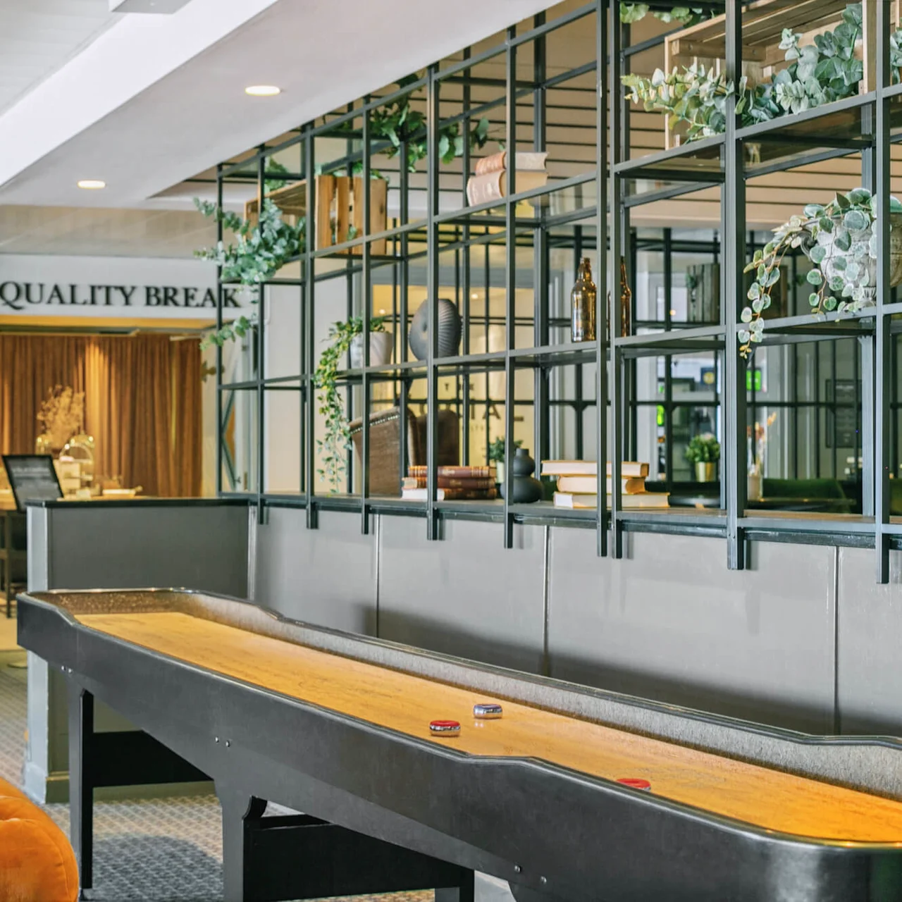 Et shuffleboardbord med pucker står i et moderne loungeområde. I nærheden er en orange sofa og dekorative hylder med planter, som skaber en hyggelig atmosfære. Tekst: "LONDON/PARIS", "QUALITY BREAK".