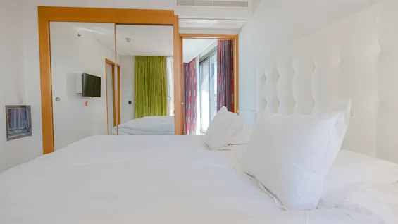 Ett ljust och modernt sovrum med en stor säng, vita sängkläder och speglade garderobsdörrar som reflekterar en TV och färgglada gardiner.