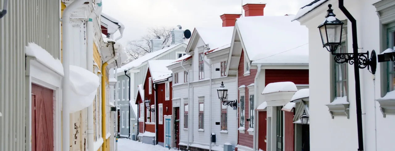 Tradisjonelle trehus langs en snødekt gate; gatelykter og snøfonner tilfører en vinterlig stemning.