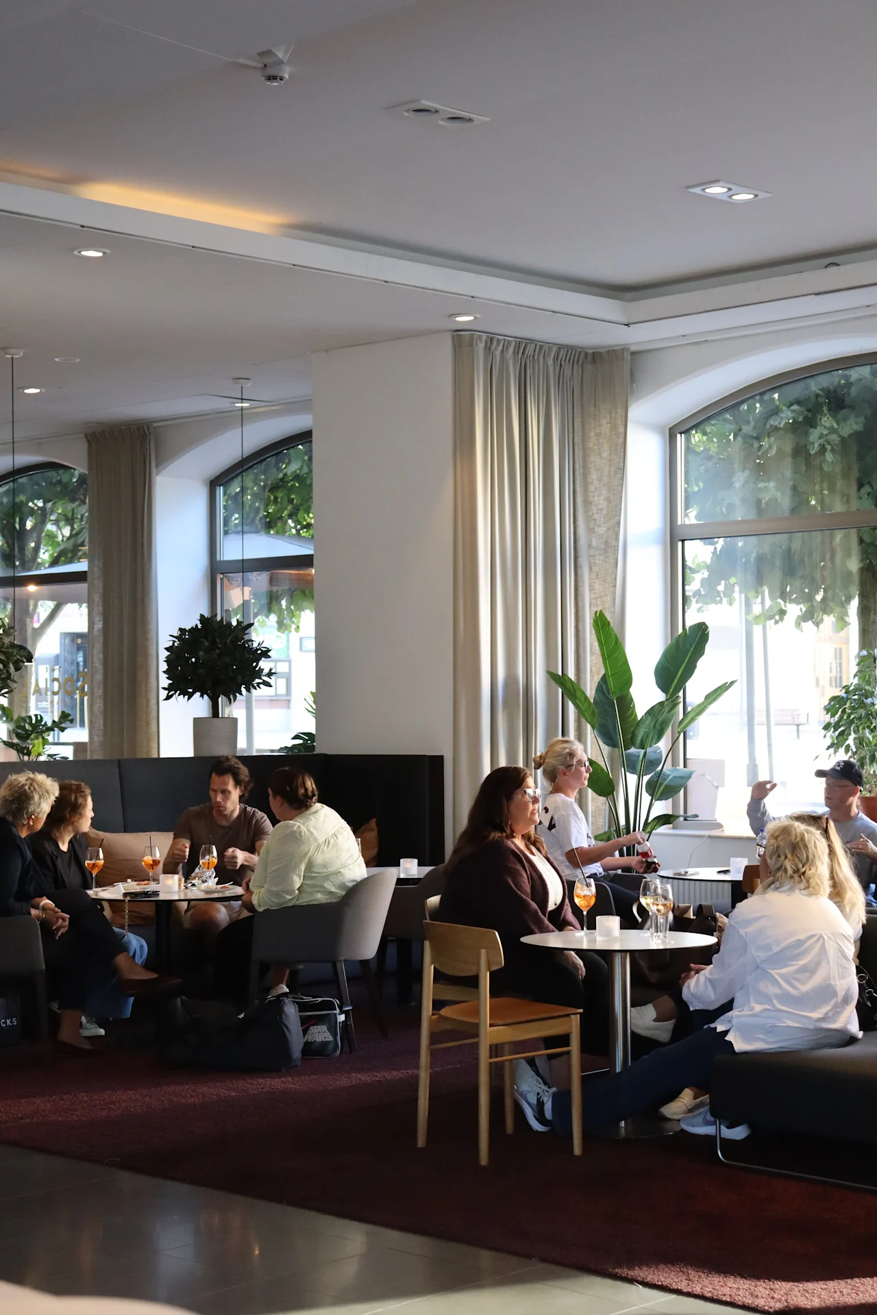 People sit and converse around tables in a warmly lit lounge area. The space features large windows, indoor plants, and modern furniture, creating a relaxed, inviting atmosphere. 