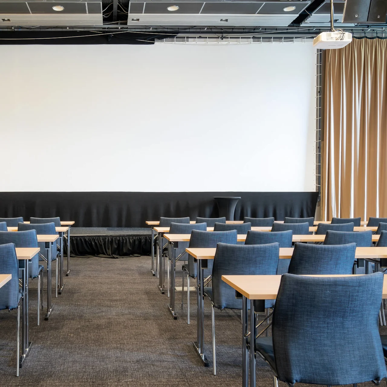 Rows of blue chairs with attached small desks in a conference room, facing a large white projection screen.