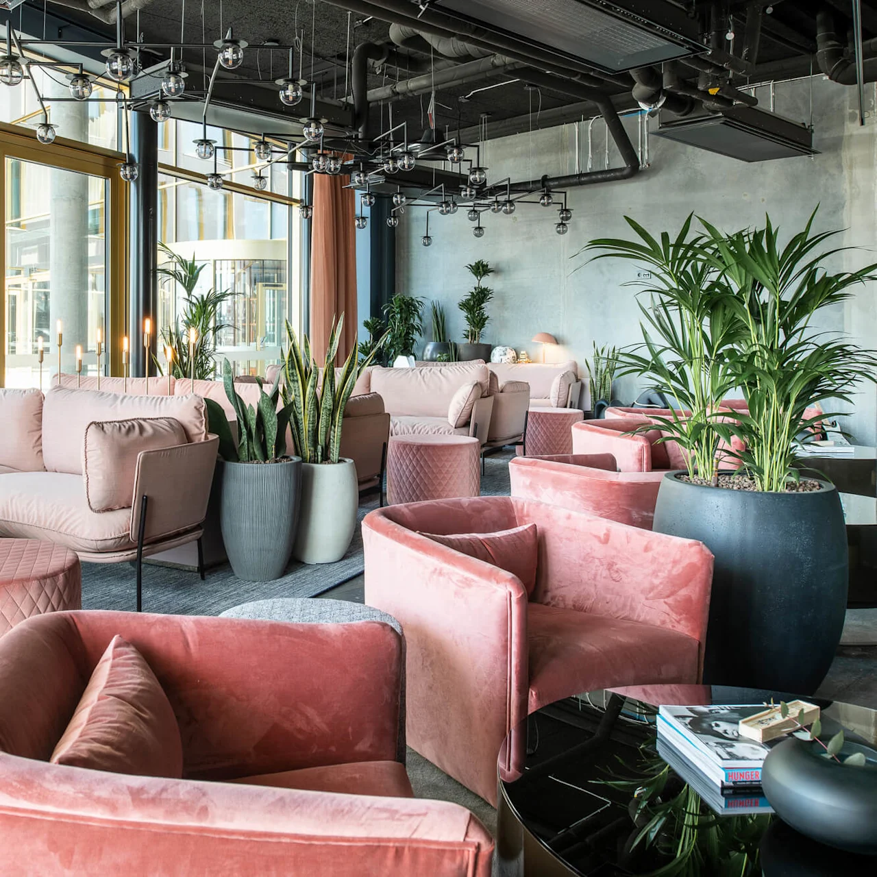Luxurious lounge with pink velvet sofas and chairs arranged around low tables. Large potted plants add greenery, set in a modern interior with floor-to-ceiling windows and a decorative ceiling.