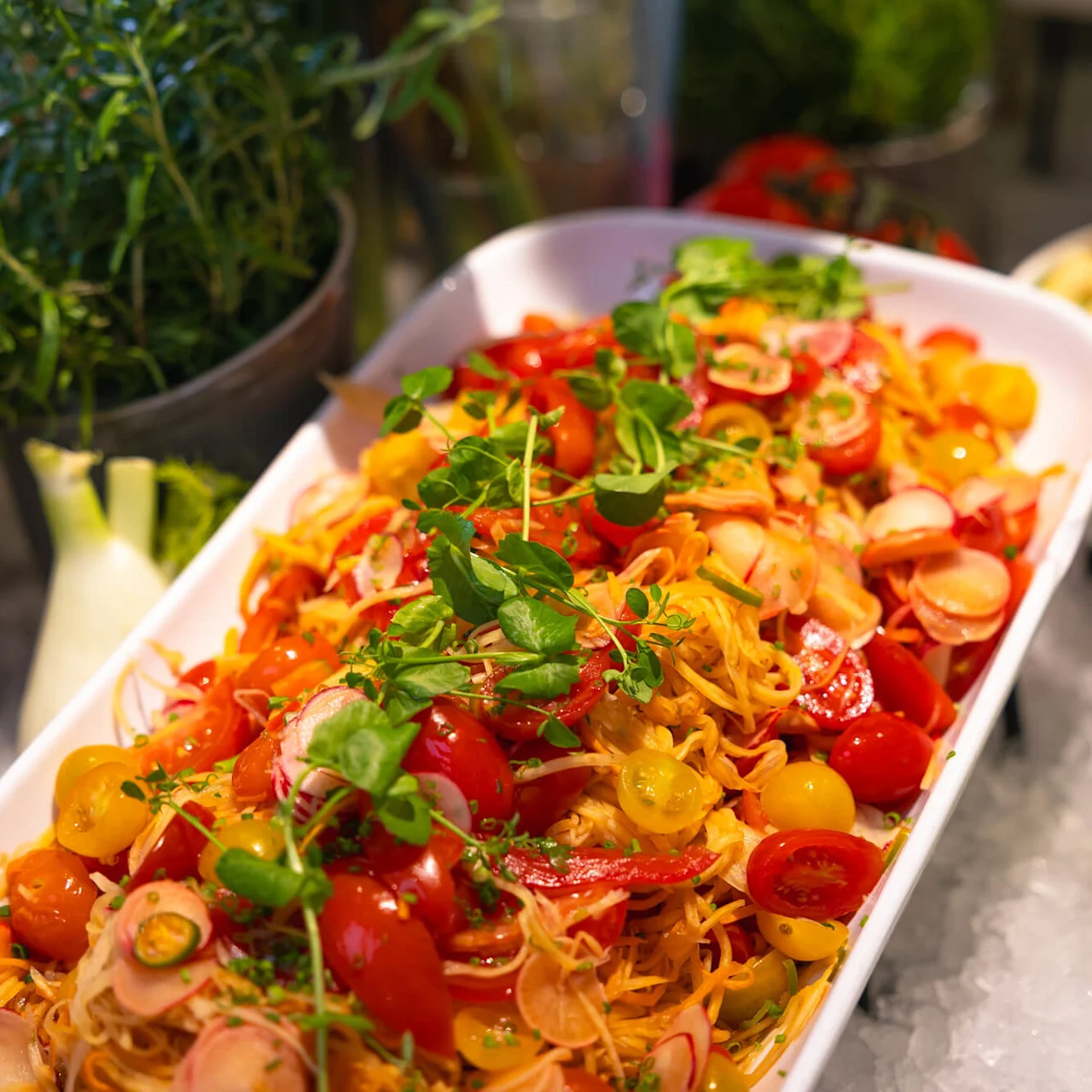 Fargerik salat med cherrytomater, reddiker og grønnsaker, arrangert i en hvit skål omgitt av urter og sitroner på et buffetbord.