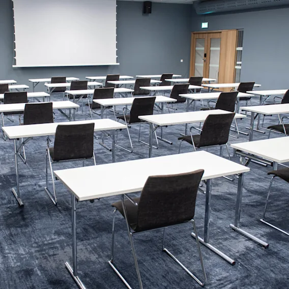 Rader av tomma skrivbord och stolar är arrangerade i ett rymligt, modernt konferensrum. Rummet har en blå matta och en stor tom skärm längst fram.