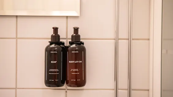 To veggmonterte flasker merket "SOAP" og "BODY LOTION" med pumper, i et flislagt bad. Teksten på hver lyder, ”MADE NOTES” og ”Hand Made”. Flaskene er brune.