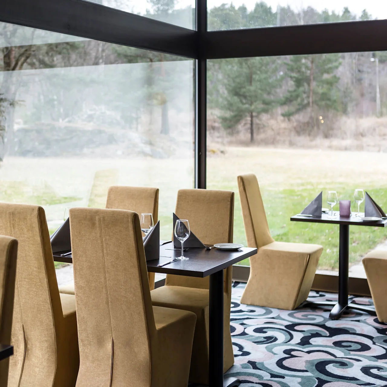 Tables are set with glasses and napkins in a restaurant with large windows overlooking a grassy landscape and trees. The chairs are beige and the carpet has a swirling pattern.