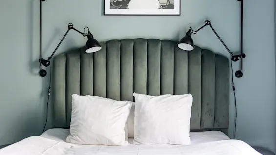 En koselig soveromsscene med en plysjgrønn sengegavl, skarpe hvite sengetøy og to stilige svarte leselamper montert på veggen.