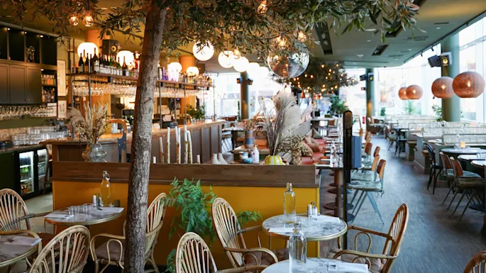 En innendørs spiseplass med bistrostoler, marmorborde, hengende lys og et tre. Atmosfæren er koselig og innbydende.