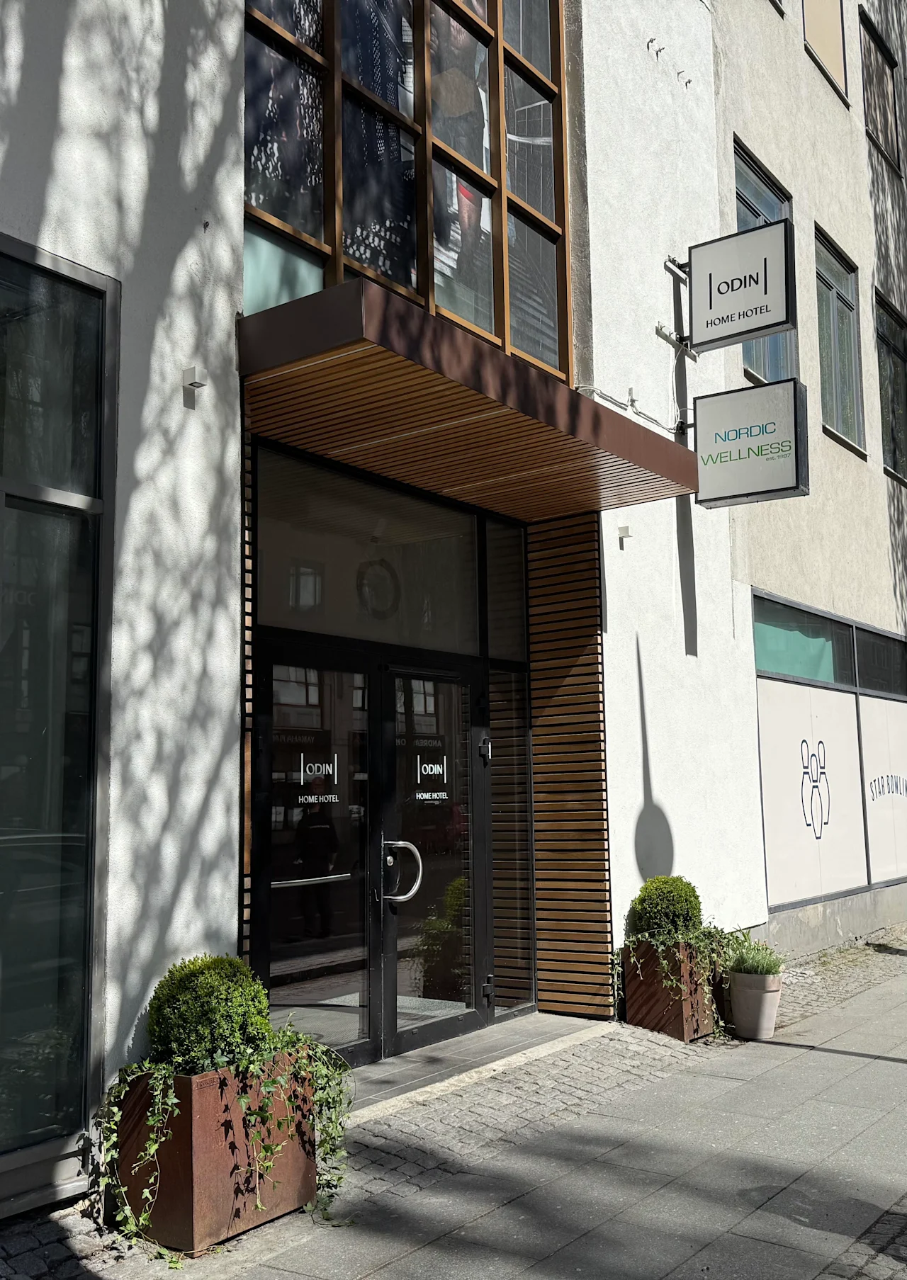 Building entrance with glass doors and a wooden canopy. Signs for HOME HOTEL and WELLNESS are visible, plus a STAR BOWLI graphic.