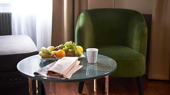 A bowl of fruit, a cup, and a newspaper on a coffee table, with a green armchair beside, in a cozy room setting.