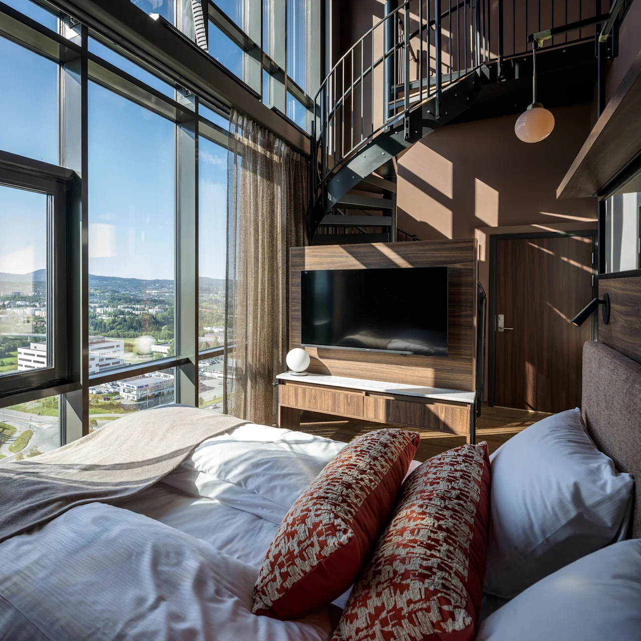 Et moderne hotelværelse med et stort vindue, der tilbyder byudsigt, en komfortabel seng, et TV og en stilfuld vindeltrappe.