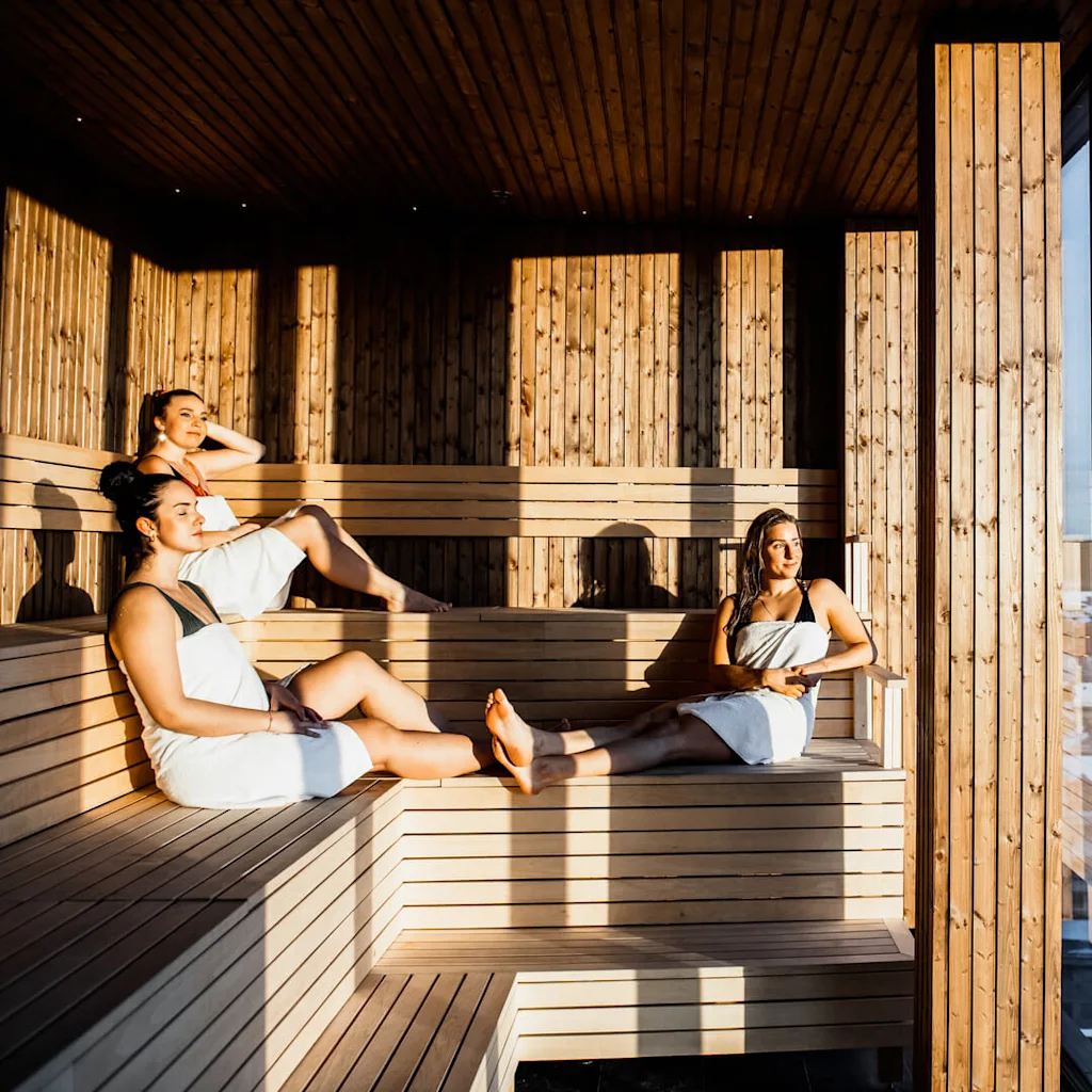 Three people are relaxing in a sunlit wooden sauna with large windows overlooking a snowy cityscape.