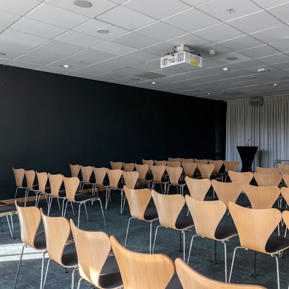 Rader av trästolar står tomma, vända mot en svart vägg och vit gardin i ett konferensrum med en takmonterad projektor.