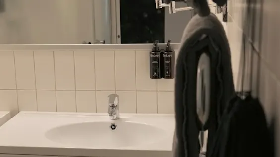En badeværelsesvask med en kromhane er omgivet af hvide fliser. To mørke flasker mærket "EcoLogiq" indeholdende håndsæbe og lotion er monteret på væggen i nærheden.