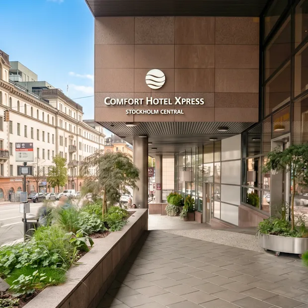 The entrance of a modern hotel in a city with green plants and a classic building in the background.