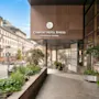 The entrance of a modern hotel in a city with green plants and a classic building in the background.