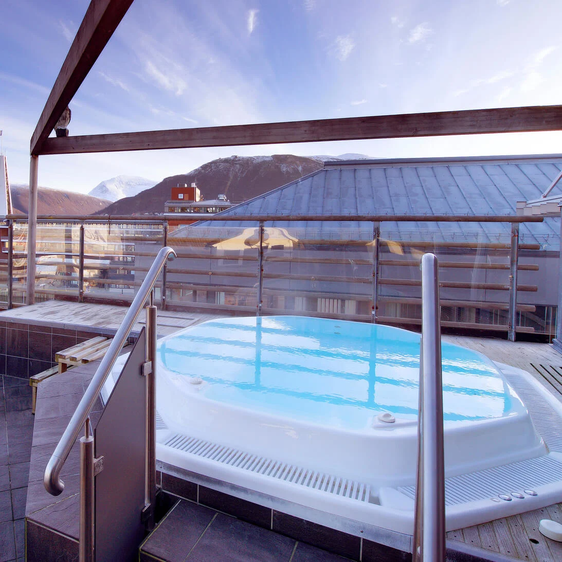 A rooftop hot tub overlooking a marina with boats, set against a backdrop of mountains and blue skies.