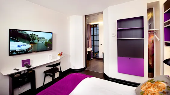 TV visar polisscen, hänger på vit hotellrumsvägg. Intill skrivbord med telefon, snacks. Lila detaljer, säng synlig. Öppen badrumsdörr avslöjar handfat, dusch.