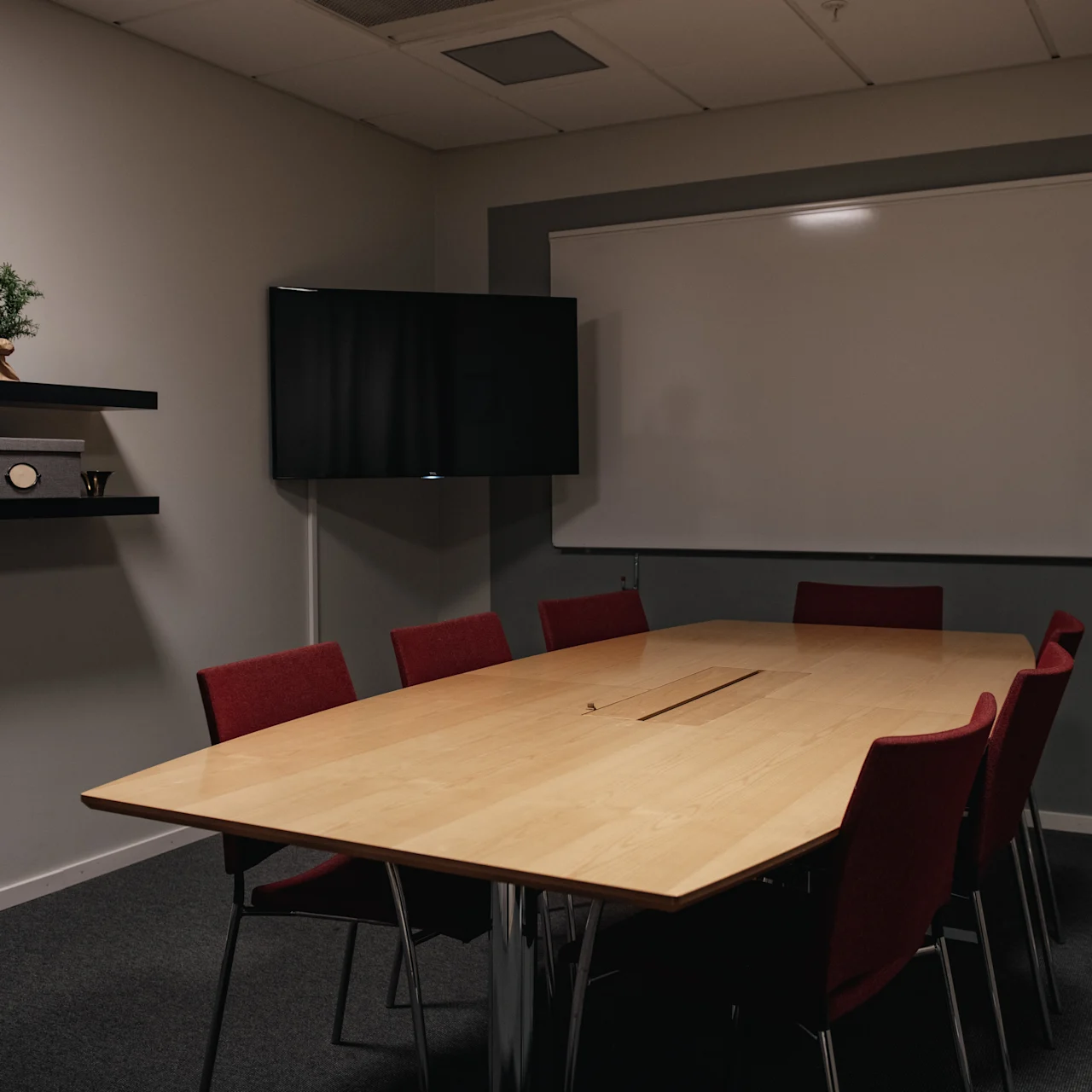 Conference table surrounded by red chairs sits in a dimly lit room. A whiteboard is mounted on the wall, with a TV screen and shelf in the background.