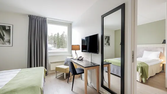 A neatly arranged hotel room features a bed with green linens and a desk beside a large mirror. The window reveals a forested view.