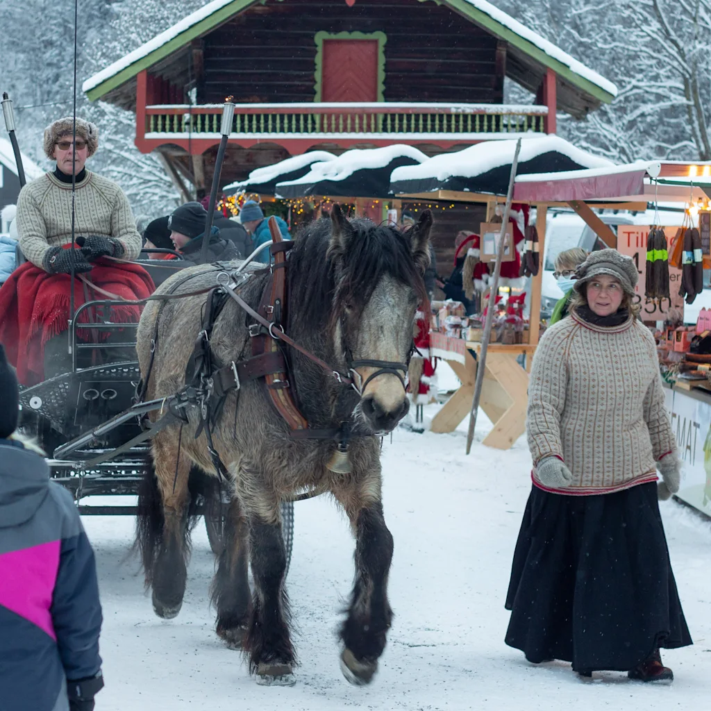 A horse-drawn carriage rides through a snowy winter market with people walking around.