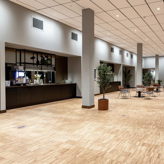 Spacious room with light wooden flooring; tables and chairs are arranged throughout. A counter with coffee machines is on one side. Potted plants are placed around the area.