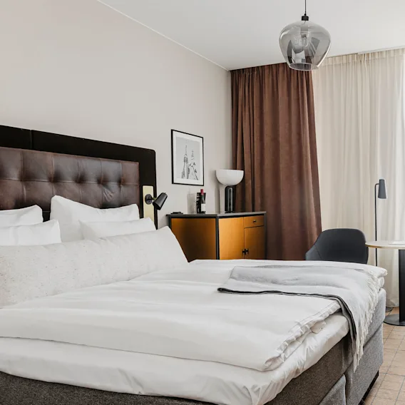 A bed with white linens and a dark headboard, situated in a modern bedroom. A lamp and artwork adorn a side table, with chairs and a table nearby, partially curtained window.
