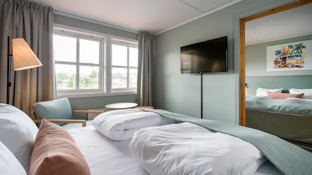 En seng med hvite og rosa puter står i et mykt opplyst rom, komplettert med en veggmontert TV og store vinduer som viser utsikten utenfor. Ved siden av er en koselig sittegruppe.