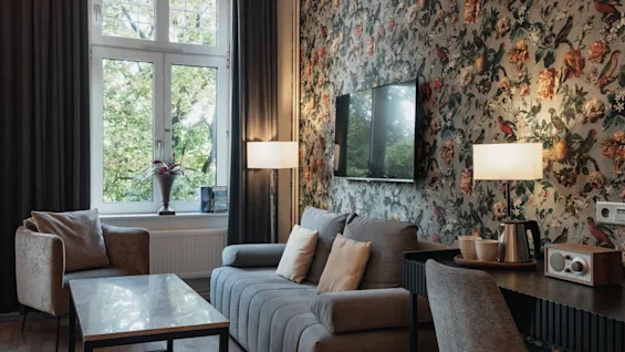 Et hyggeligt hotelværelse på Home Hotel Norre Park i Halmstad med en mønstret væg, sofa, lænestol og et skrivebordsområde med en elkedel.