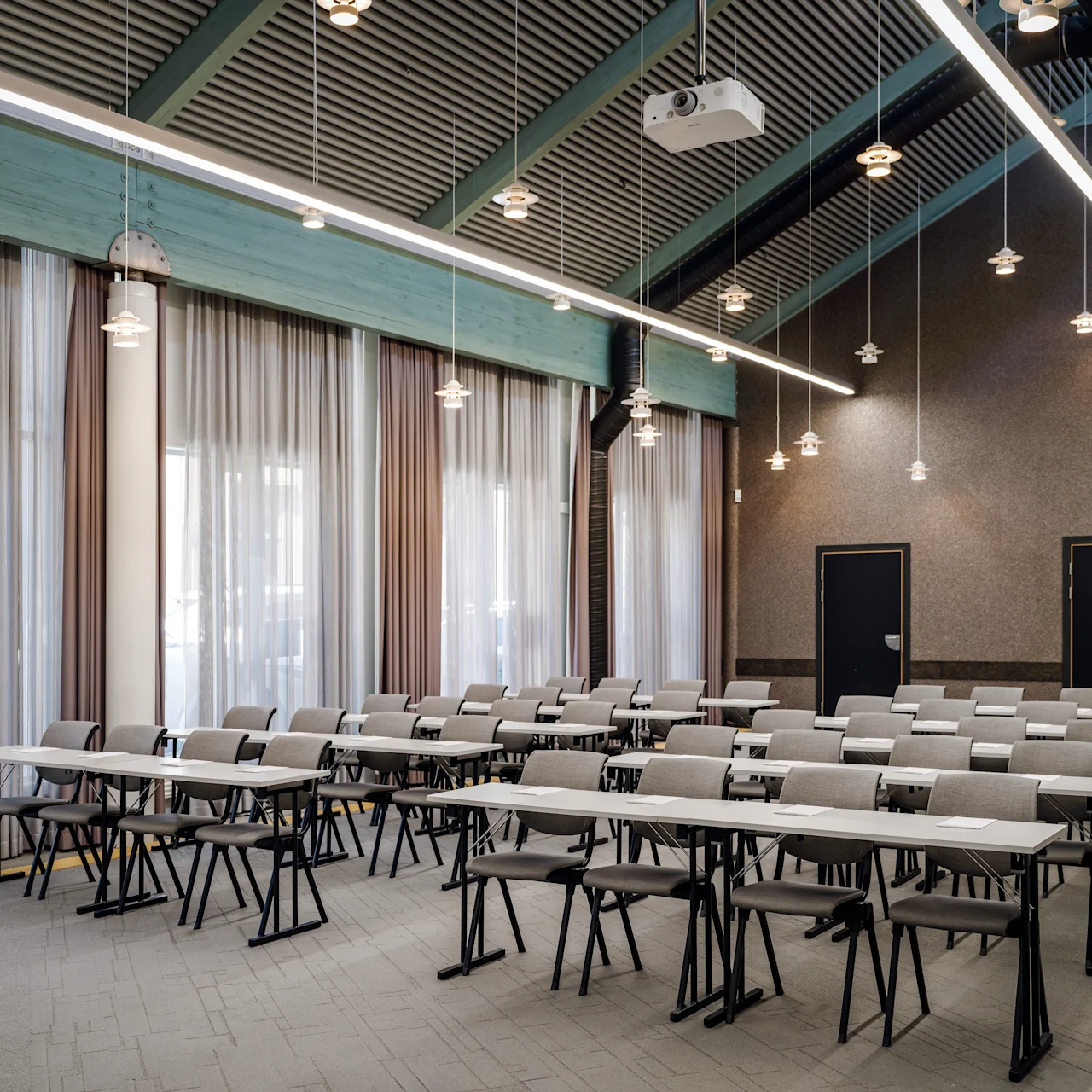 Rows of empty desks with chairs arranged in a conference room; overhead lights and a projector hang from the high ceiling. Curved curtains cover large windows on one wall.
