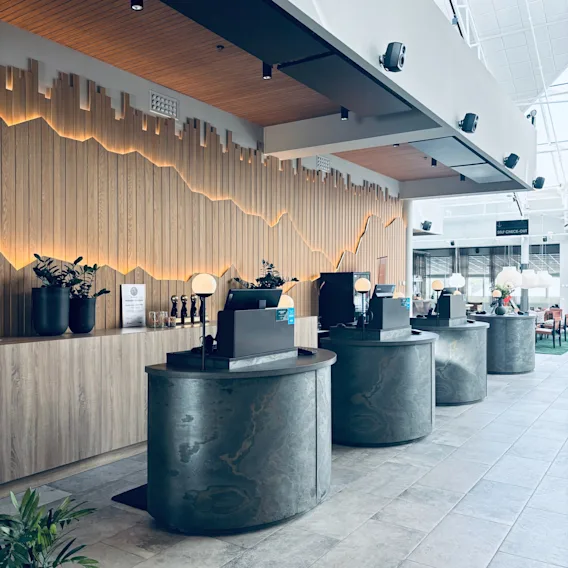 En modern hotellobby med flera självbetjäningsstationer för incheckning. En dekorativ trävägg med bakgrundsbelysning ger värme. SELF CHECK-OUT.