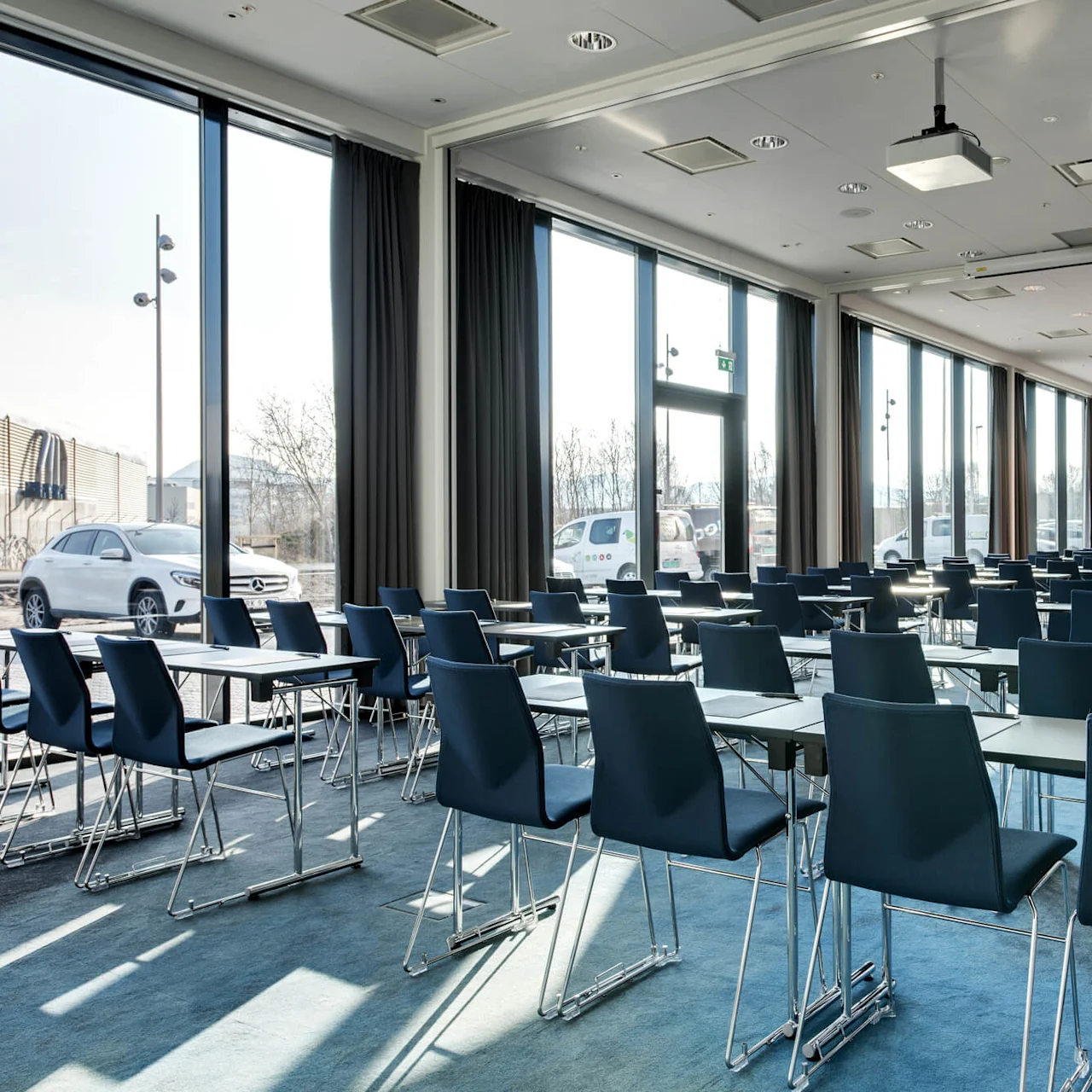 Rows of empty navy chairs face a large white screen in a bright conference room with floor-to-ceiling windows and blue carpet. A Mercedes-Benz car is visible outside.