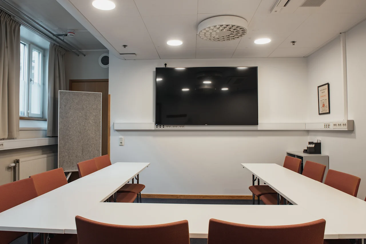 A bright conference room at Home Hotel Uppsala featuring a U-shaped table arrangement, comfortable chairs, and a large display.