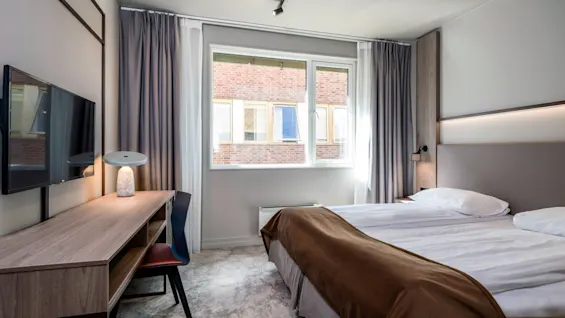Et hotelværelse med en redt dobbeltseng, en vægmonteret TV og et skrivebord med en lampe. Stort vindue med udsigt til murstensbygning.